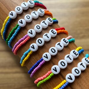 May include: Several colorful beaded bracelets spelling "VOTE" are displayed. Each bracelet features white letter beads and a rainbow of colors. The bracelets are arranged on a wooden surface, showcasing their vibrant design.