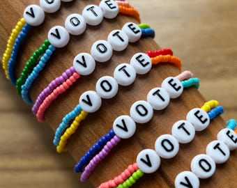Pode incluir: Várias pulseiras coloridas com a palavra "VOTE". Cada pulseira apresenta contas de letras brancas e um arco-íris de cores. As pulseiras estão dispostas sobre uma superfície de madeira, mostrando seu design vibrante.