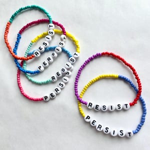 May include: Five colorful beaded bracelets with the words "Resist" and "Persist" spelled out in white letters.