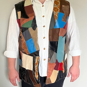 Puede incluir: Un chaleco sin mangas confeccionado con un mosaico de piezas de cuero y ante multicolor en tonos negro, marrón, azul, rojo, verde y crema, usado sobre una camisa blanca abotonada con mangas remangadas y pantalones oscuros.
