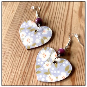 May include: A pair of heart-shaped earrings with a floral pattern on a light purple background. The earrings have silver hooks and a purple gemstone bead.