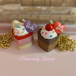 Puede incluir: Dos colgantes de pastel en forma de corazón con detalles de cadena dorada. Uno es rosa y amarillo con un corazón morado que dice "Barbie". El otro es marrón con una cereza roja y un corazón rojo que dice "Barbie". El texto "Insanely Sweet" está en la parte inferior.