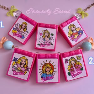 以下が含まれることがあります： バービーをテーマにした6つのピンク色のペンダントが付いたネックレス。それぞれ異なるバービーのイラストが描かれています。ペンダントは、リボンやビーズなどの装飾要素が付いたゴールドチェーンで繋がれています。背景は薄いピンク色です。