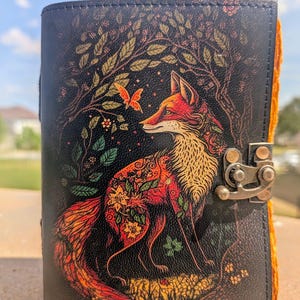 May include: A black leather journal with a colorful illustration of a fox standing in a forest. The fox is surrounded by flowers and leaves, and there is a butterfly flying above it. The journal has a brass clasp.