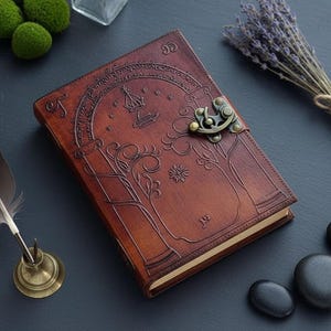 Puede incluir: Un diario de cuero marr&oacute;n con un dise&ntilde;o en relieve de &aacute;rbol y corona, asegurado con un cierre met&aacute;lico. La imagen incluye una pluma, lavanda y piedras. El diario est&aacute; cerrado.
