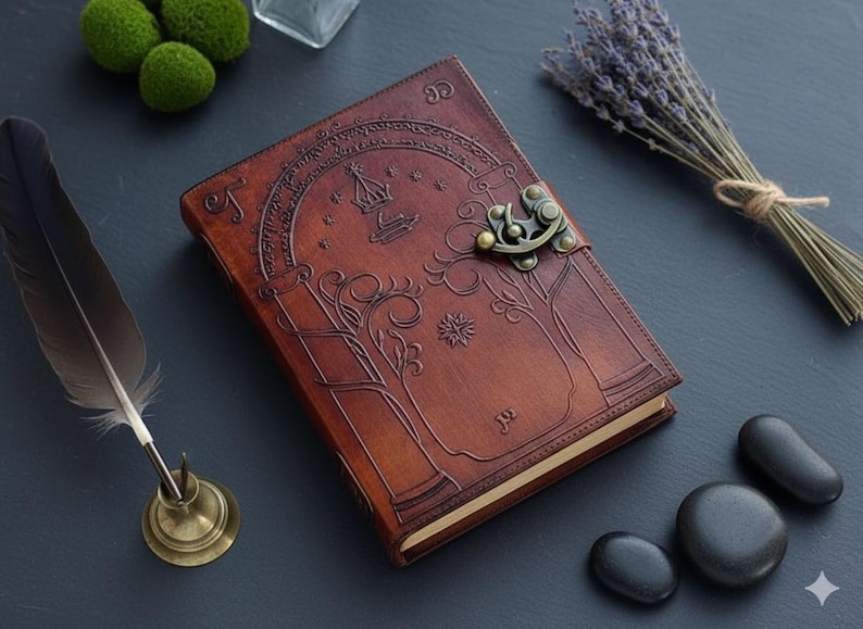 Puede incluir: Un diario de cuero marr&oacute;n con un dise&ntilde;o en relieve de &aacute;rbol y corona, asegurado con un cierre met&aacute;lico. La imagen incluye una pluma, lavanda y piedras. El diario est&aacute; cerrado.