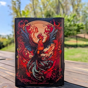 May include: Black leather wallet featuring a vibrant phoenix design. The artwork showcases a fiery bird with red, orange, and blue plumage against a backdrop of flames and a setting sun. The wallet has a snap closure and is displayed on a wooden surface.