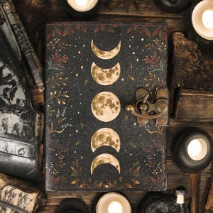 Puede incluir: Un diario de cuero negro con un cierre dorado y un dise&ntilde;o de fases lunares. El diario est&aacute; decorado con detalles florales dorados y marrones y un fondo de noche estrellada.
