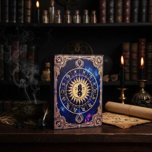 Puede incluir: Un libro de tapa dura con un dise&ntilde;o celestial. La portada presenta un fondo azul con una ilustraci&oacute;n del sol y la luna rodeada de s&iacute;mbolos del zodiaco. El libro est&aacute; sobre una superficie de madera con velas y libros en el fondo.