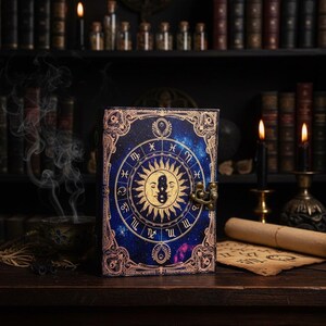 Puede incluir: Un diario azul oscuro con un dise&ntilde;o celestial que presenta s&iacute;mbolos del zod&iacute;aco y una ilustraci&oacute;n del sol y la luna. La portada tiene un borde decorativo y un cierre de metal. El diario est&aacute; sobre una superficie de madera con velas y libros en el fondo.