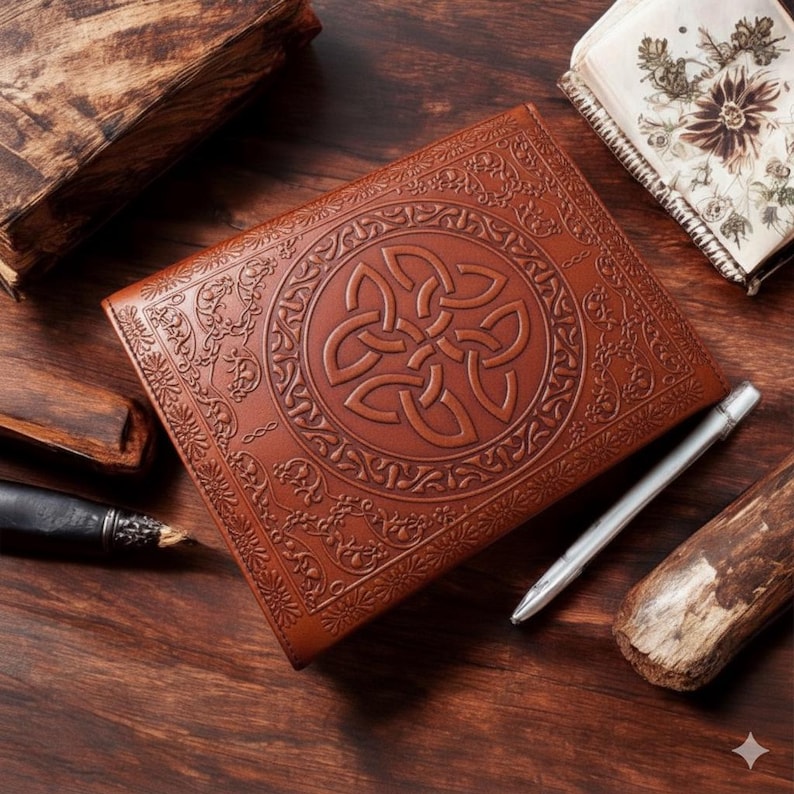 Puede incluir: Un diario de cuero marr&oacute;n con un dise&ntilde;o de nudo celta en relieve. Un bol&iacute;grafo plateado y otros libros est&aacute;n sobre una superficie de madera. El diario est&aacute; abierto, revelando p&aacute;ginas en blanco.
