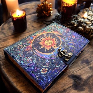 May include: A decorative journal with a celestial design featuring a sun and moon on the cover. The cover is adorned with intricate floral patterns in shades of blue, purple, and orange. It has a black leather strap with a metal clasp. Candles are in the background.