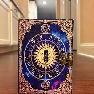 Puede incluir: Un diario azul oscuro con un dise&ntilde;o celestial que presenta s&iacute;mbolos del zod&iacute;aco, un sol y una luna. La cubierta tiene un borde dorado y un cierre de metal. El diario est&aacute; de pie sobre una superficie de madera.