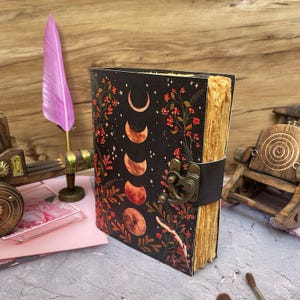 Puede incluir: Un diario de cuero negro con un dise&ntilde;o floral y lunar. El diario tiene un cierre de broche y un aspecto desgastado y envejecido.