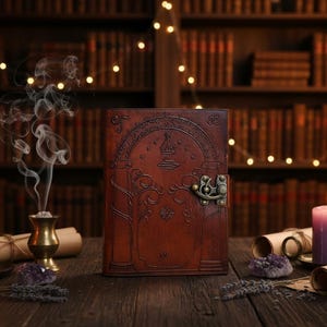 Puede incluir: Un diario de cuero marr&oacute;n con un dise&ntilde;o en relieve que presenta una puerta arqueada, &aacute;rboles y estrellas. El diario tiene un cierre de metal y est&aacute; rodeado de velas, pergaminos y cristales de amatista. El fondo incluye una estanter&iacute;a y luces de hadas.