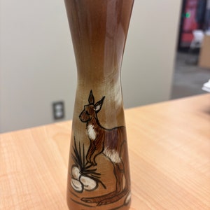 May include: A brown ceramic vase with a kangaroo design. The vase has a narrow neck and a wide base. The kangaroo is brown with white markings and is standing on its hind legs. There are also some white eggs and a brown plant in the design.