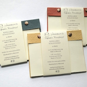 Op de afbeelding: Verschillende navulbare krasblokken met ivoorkleurig papier worden getoond. Elk blok heeft een gekleurde kaft in groen, beige en rood, vastgezet met een messing schroef. De blokken bevatten de tekst "HANDBOUND" en "REFILLABLE SCRATCH PAD".