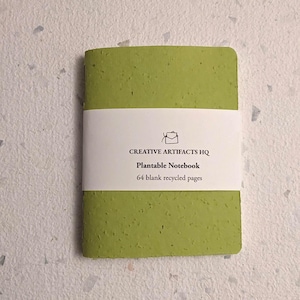 May include: A light green notebook with a textured, speckled cover. A white paper band wraps around the centre, featuring the text "CREATIVE ARTIFACTS HQ", "Plantable Notebook", and "64 blank recycled pages" with a small envelope icon.