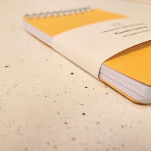 May include: A yellow spiral-bound notebook with a white paper band wrapped around the center. The band features printed text that reads Creative Articulate Notebook and 64 blank recycled pages. The notebook has white pages and a yellow cover.