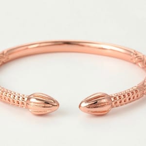 May include: Rose gold open cuff bracelet. The bracelet has a textured design along the band and at each end. The ends of the bracelet are shaped like a seed or a bud. The bracelet is shown against a white background.