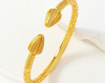 May include: A gold-colored cuff bracelet with a snake-like design. The bracelet features textured detailing and two stylized snake heads at the ends. The bracelet is made of a shiny, reflective metal.