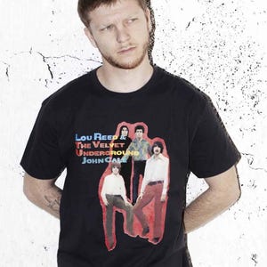 May include: Black crew neck t-shirt featuring a graphic of Lou Reed & The Velvet Underground and John Cale. The graphic is a vintage-style image of the band members in a red border. The text is in blue, red, and yellow.