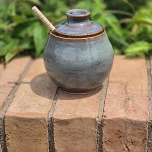 May include: A round, blue-grey ceramic honey pot with a matching lid and a light-colored wooden honey dipper inserted into the top opening. The pot features a smooth, glossy glaze with subtle variations in color and a dark brown base rim.