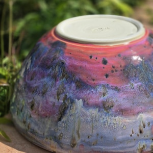 May include: A ceramic bowl featuring a vibrant glaze with swirling patterns of pink, purple, and blue. The exterior displays a mottled texture with dark speckles, while the circular base is unglazed, showing a light beige clay with a faint signature.