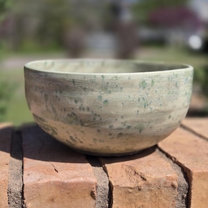 May include: A green and white ceramic bowl with a speckled glaze. The bowl is sitting on a brick surface.