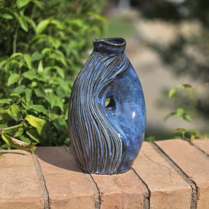 May include: A ceramic vase with a deep blue glaze and a glossy finish. The vessel features a unique sculptural form with a central cutout and a side section decorated with vertical, textured grooves that reveal dark brown clay beneath the blue glaze.