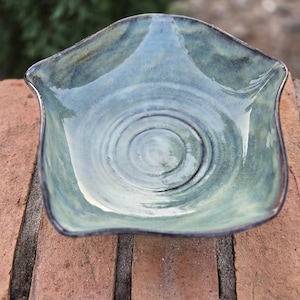 May include: A handmade, square ceramic bowl with a wavy rim. The bowl has a glossy, blue-green glaze with darker blue and brown accents. The interior features concentric circles, and the rim is outlined in dark brown.
