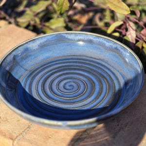 May include: A handmade ceramic bowl with a spiral design in shades of blue and brown. The bowl has a glossy finish and a slightly uneven rim. The bowl is sitting on a brick surface.