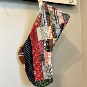 May include: A handmade Christmas stocking with a patchwork design. The stocking is made from various fabric scraps in red, blue, green, and white. The stocking is hanging on a wall.