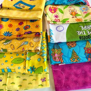 May include: A collection of colorful fabric pieces with various patterns. Designs include cartoon characters, flowers, and whimsical illustrations in yellow, blue, and pink. Some fabrics have text. Ideal for quilting or crafting projects.