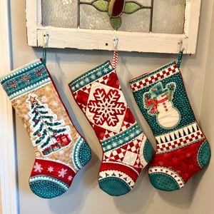 May include: Three quilted Christmas stockings featuring festive patterns. The left stocking displays a decorated tree, the center features a large white snowflake on a red background, and the right shows a snowman wearing a hat and scarf.