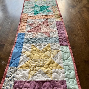 May include: A patchwork table runner with a variety of colors and patterns, including a yellow star, a pink and white geometric design, and a blue floral print.