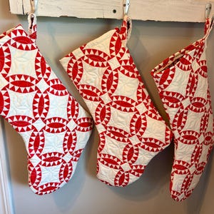 May include: Three quilted Christmas stockings featuring a repeating red and white pattern of interlocking circles and geometric shapes. Each stocking has a fabric loop at the top for hanging and displays detailed stitching across the surface.