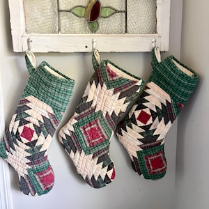 May include: Three quilted Christmas stockings hanging from a white wooden frame. Each stocking features a geometric patchwork design in shades of green, cream, and dark red, constructed from various plaid and solid fabric squares with visible stitching.