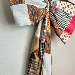 May include: A large, handmade fabric bow with a patchwork design. The bow features a variety of patterns and colors, including paisley, floral, and gingham. The bow is tied in the center and hangs vertically.