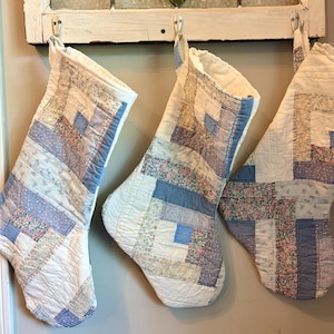 May include: Three quilted Christmas stockings hanging in a row, each featuring a patchwork design of blue, white, and floral fabric strips. The stockings are constructed with a log cabin pattern and have a white fabric cuff at the top opening.