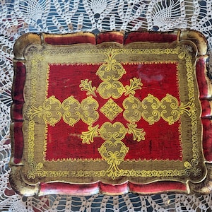 May include: Rectangular decorative tray with a red center and gold trim. The tray features ornate gold scrollwork and a scalloped edge. The tray's frame is a mix of gold and red, creating a vintage aesthetic.