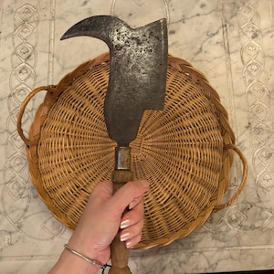 May include: A large, curved, metal blade with a wooden handle. The blade is rusty and has a worn appearance. The blade is resting on a woven wicker basket.