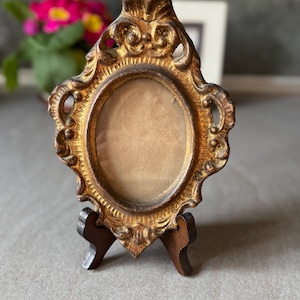 Könnte beinhalten: Ein kleiner, verzierter goldfarbener Bilderrahmen mit einer ovalen Öffnung. Der Rahmen hat ein dekoratives Design und steht auf einem kleinen Holzständer.