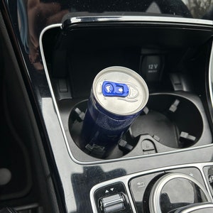 Könnte beinhalten: Eine blaue und silberne Energy-Drink-Dose in einem Getränkehalter im Auto. Die Dose hat ein blaues und silbernes Etikett. Der Innenraum des Autos ist schwarz mit silbernen Akzenten, einschließlich Armaturenbrett.