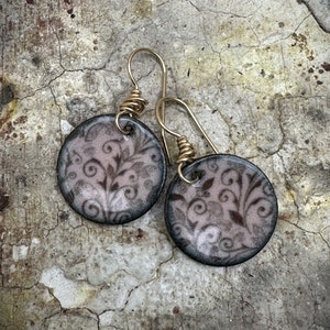 May include: A pair of round ceramic earrings with a delicate floral pattern in shades of pink and black. The earrings have gold-toned wire hooks.