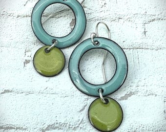 May include: A pair of handmade earrings featuring a teal enamel ring with a small olive green disc pendant. The earrings have silver-colored ear wires. The jewelry is set against a white brick background.