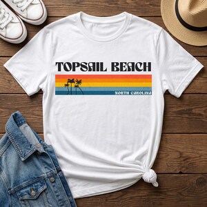 May include: White t-shirt with the text "TOPSAIL BEACH NORTH CAROLINA" in black. The design includes a horizontal stripe with orange, yellow, and blue colors, and palm tree silhouettes. The shirt is on a wooden surface.