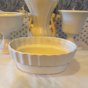 Op de afbeelding: Een verzameling witte keramische vazen en een gegroefde schaal. De schaal is ovaal met een geschulpte rand en een lichtgele binnenkant. De vazen hebben verschillende vormen, waaronder een met handvatten en een op een voetstuk.