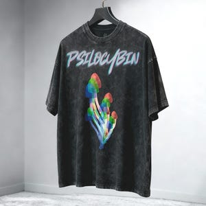 Op de afbeelding: Donkergrijs t-shirt met het woord "PSILOCYBIN" in een blauw en wit lettertype. Daaronder een kleurrijke afbeelding van paddenstoelen. Het t-shirt hangt aan een zwarte hanger.