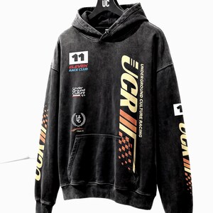 May include: Black hooded sweatshirt with "UCR" and "Underground Culture Racing" text in gold and orange. Features patches with "11 Eleven Race Club" and a logo. The hoodie has a front pocket and ribbed cuffs.